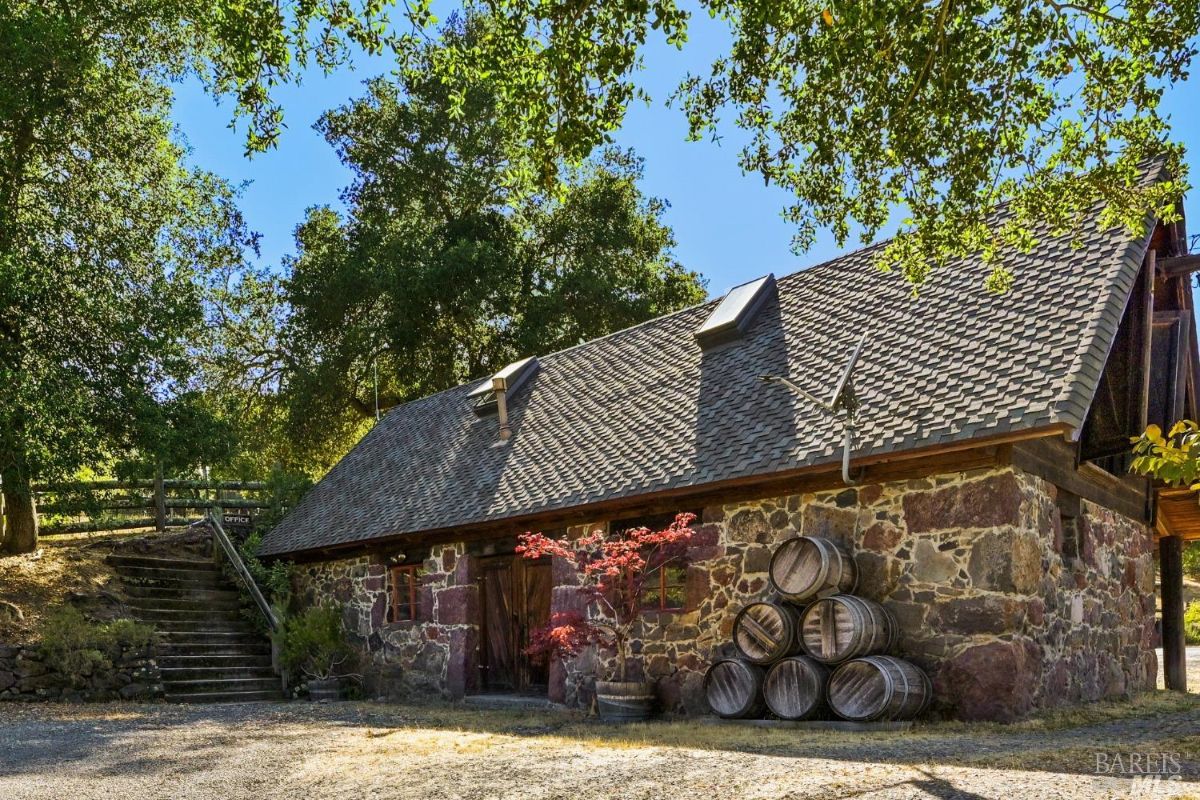 Stone building with a steep, shingled roof and two skylights, surrounded by trees. A stack of wooden barrels is arranged beside the building, and a small red-leafed tree grows near the entrance. Stone steps to the left lead up to a wooden fence, and a gravel area stretches out in the foreground.