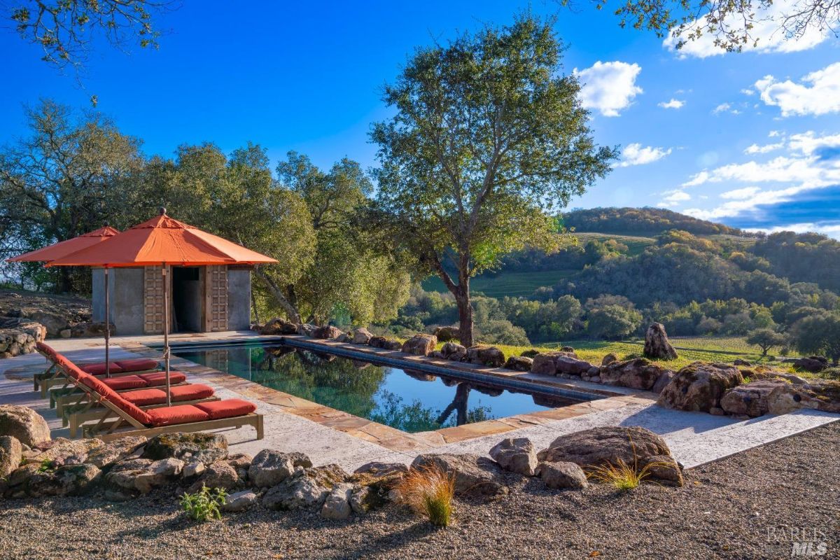 Outdoor swimming pool surrounded by a stone deck with red lounge chairs and matching umbrellas. A small structure with wooden double doors is located at the far end of the pool. The background features trees, hills, and a clear blue sky under natural daylight.