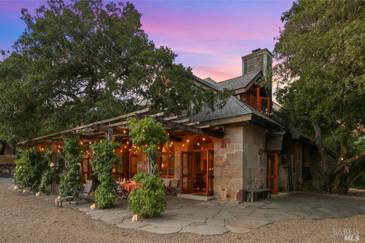 Stone house with a pergola extending over an outdoor seating area, surrounded by greenery and string lights. A large tree with sprawling branches partially covers the house, and the windows emit a warm light. The setting features a gravel ground, stone patio, and a soft-colored sunset sky in the background.