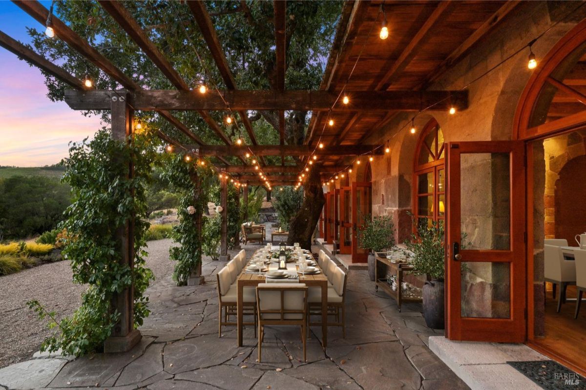 Outdoor dining area on a stone patio with a wooden pergola overhead. The pergola has string lights hanging across its beams, and climbing plants grow along its wooden posts. A long dining table with white chairs is set for a meal, including tableware and centerpieces. To the right, there are open wooden doors leading into a building with arched windows and stone walls. The background features trees, plants, and a gravel pathway.