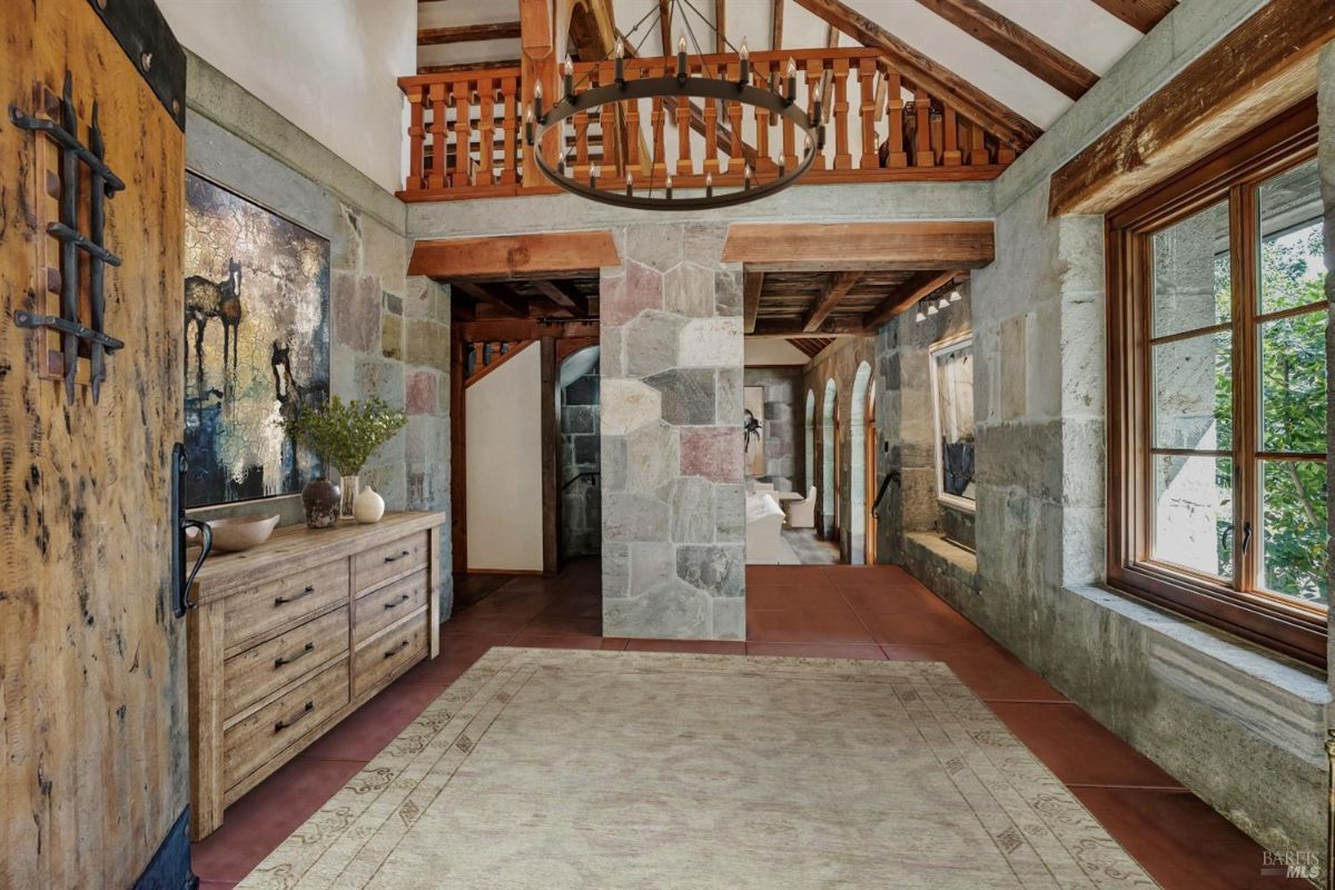Entryway with stone walls, wooden beams on the ceiling, and a large chandelier hanging above. A wooden dresser with drawers is positioned against one wall, topped with decorative items, including a vase and a bowl. A hallway leads further into the space, with large arched windows and doorways visible in the background, allowing natural light to enter.