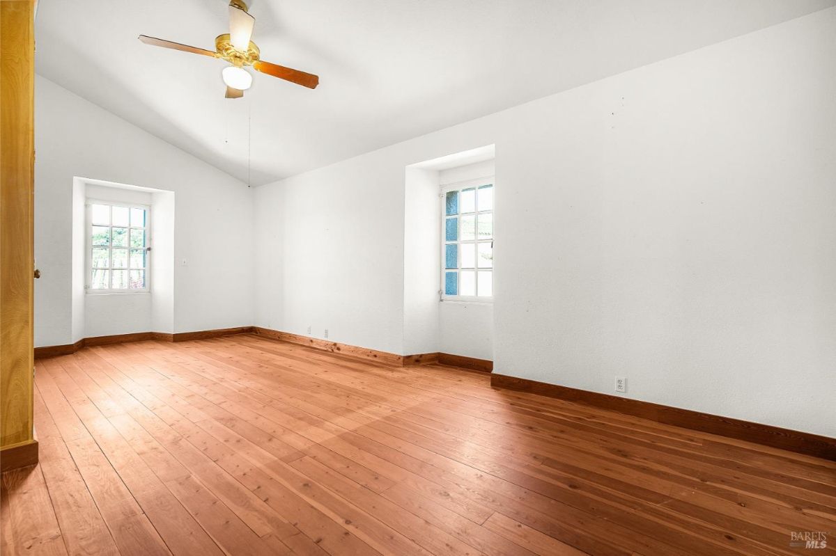 A room with wood floors, white walls, a ceiling fan, and two windows letting in natural light.