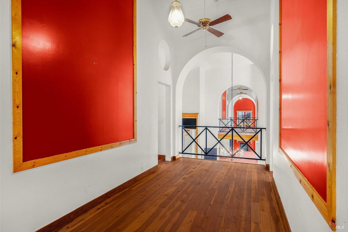 A hallway with wood floors, red accent panels, arched doorways, and a railing overlooking a lower level.