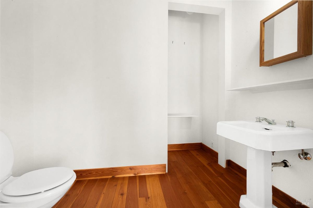 A small bathroom with wood flooring, a white pedestal sink, and a toilet against plain walls.