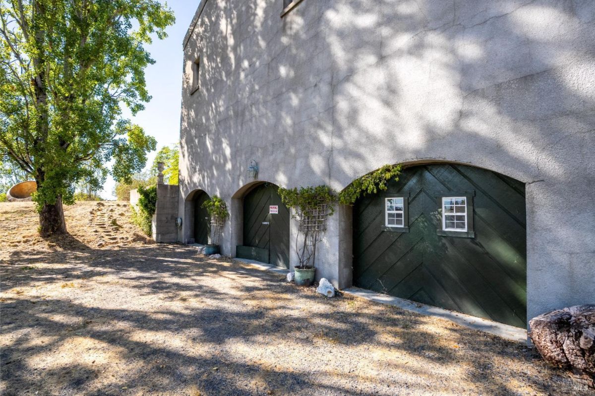 A building exterior with arched green doors and potted plants along a shaded gravel pathway.