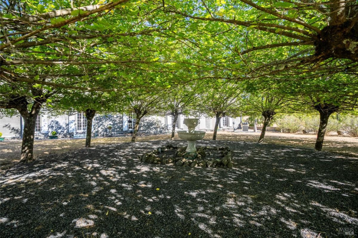 Fountain surrounded by trees in a shaded courtyard with gravel flooring.