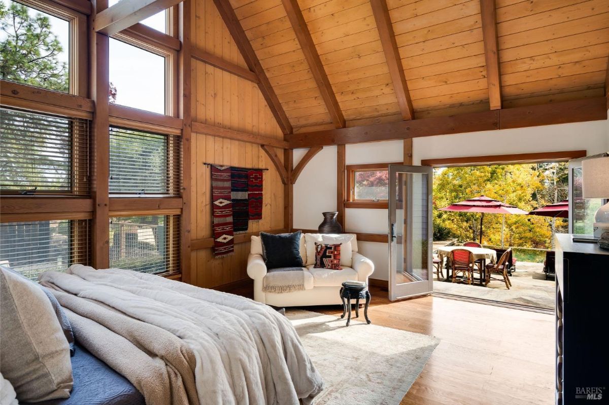 Bedroom with vaulted wood ceilings, a large bed, and doors opening to a patio with outdoor seating and umbrellas.