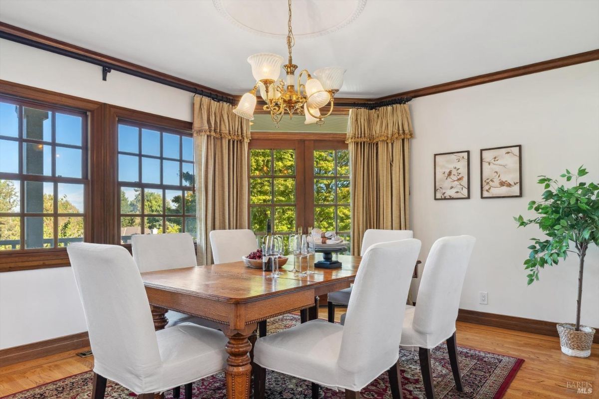 A formal dining room with a large wooden table, white chairs, and a chandelier and has large windows with views of the outdoors.
