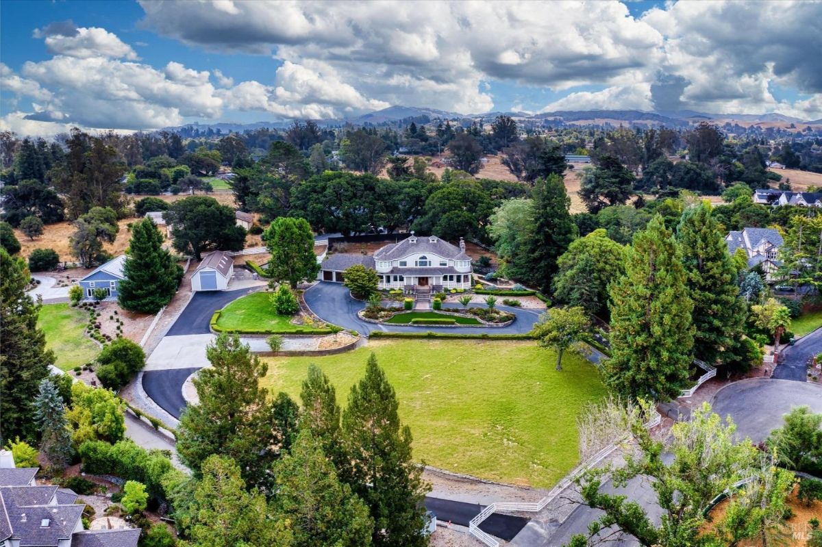 Aerial view of a large house with a circular driveway, landscaping, and surrounding trees, with distant hills visible in the background.
