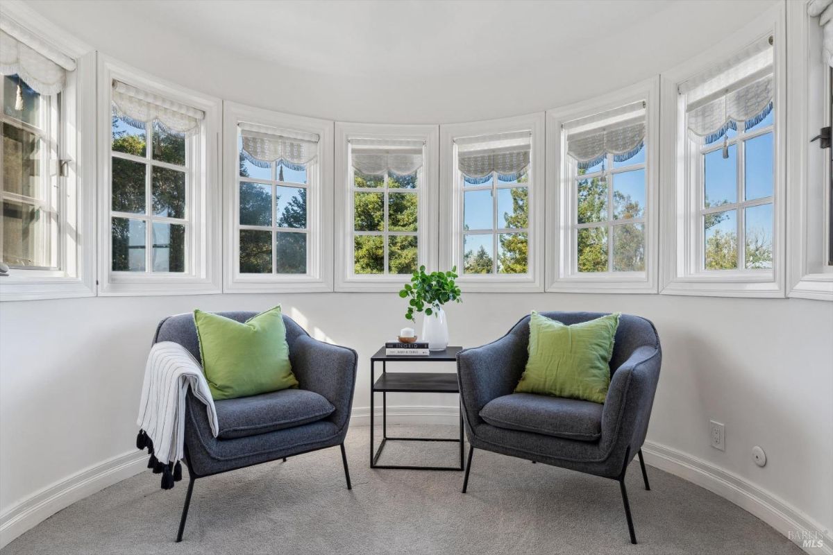 Cozy reading nook or sitting area, it has a circular shape with multiple windows offering views of the outdoors and two comfortable armchairs with green pillows and a small table with a vase and book create a relaxing atmosphere.