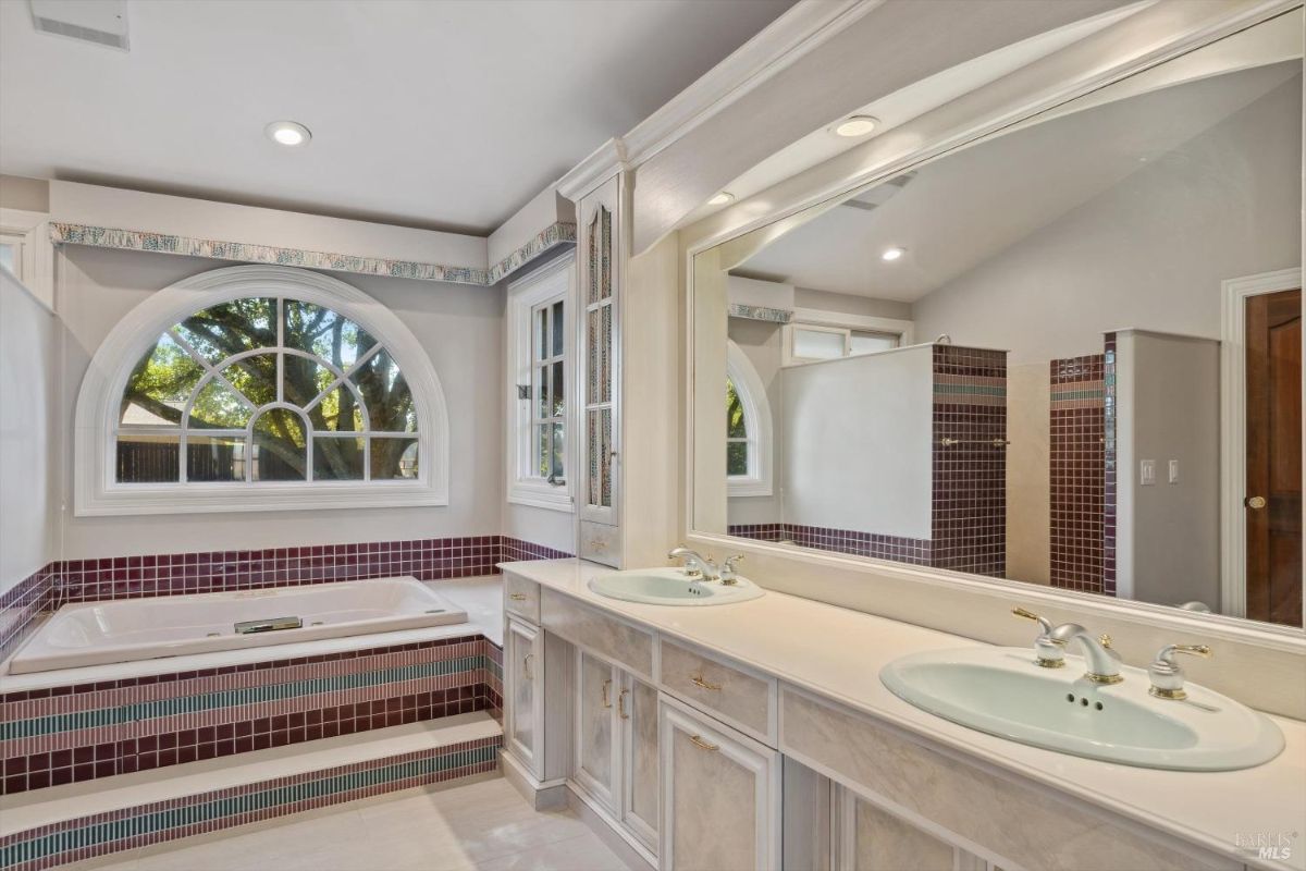 Bathroom with dual sinks, a large mirror, and a raised bathtub surrounded by purple and teal tiles.