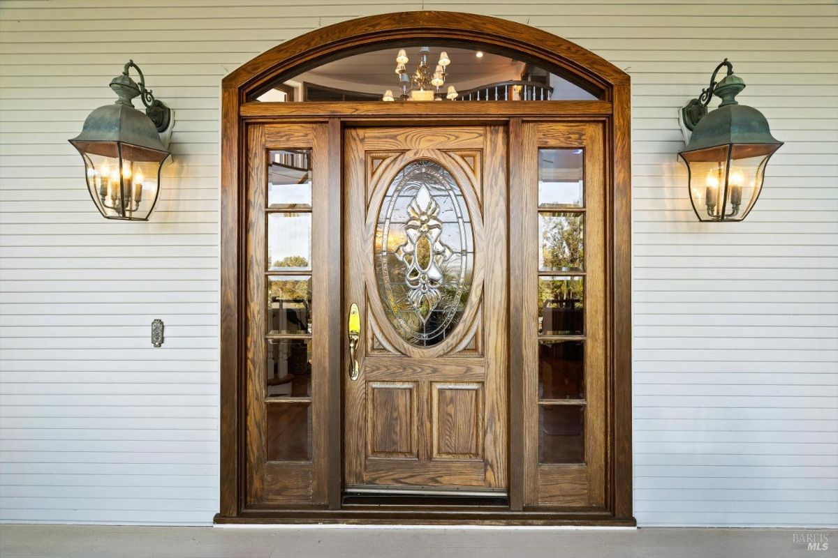 Front entrance of a house with a door made of wood with a stained glass window and decorative metal hardware and there are two light fixtures on either side of the door.