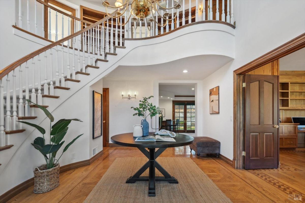 Interior of the home with the entryway features a grand staircase with a curved banister, hardwood floors, a round table with a vase and book, a plant in a basket, and a door leading to another room.