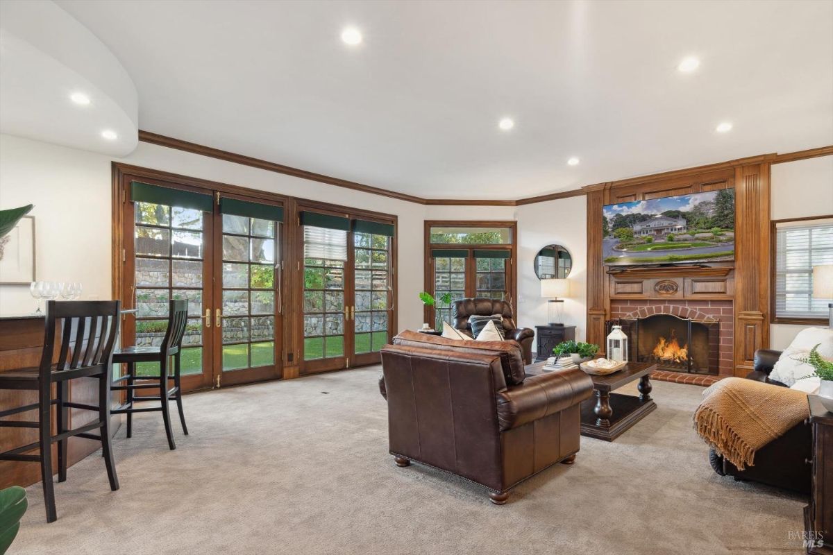 Living room with a fireplace, a large TV mounted above it, and a seating area with leather couches and a coffee table, also a bar area with stools and a window leading to the outside. 