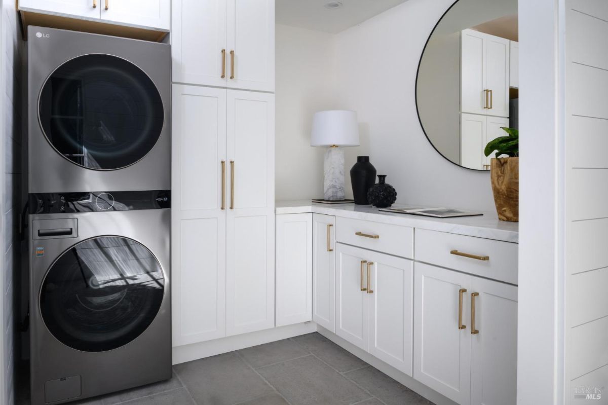 Laundry room with white cabinets, a stacked washer and dryer, and a countertop with a round mirror above it.