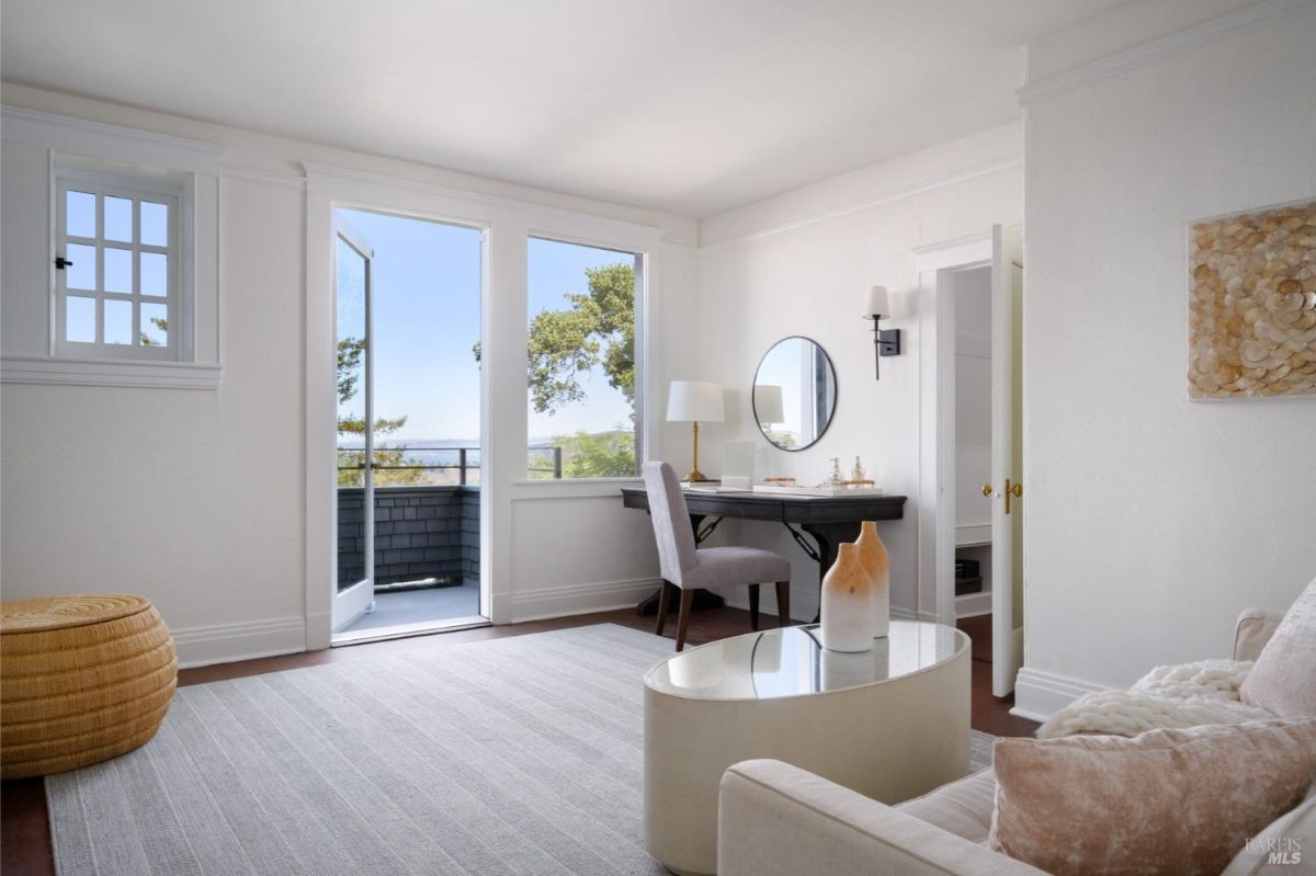 A bedroom or sitting room with white walls, a door leading to a balcony, and a view of a garden or park.