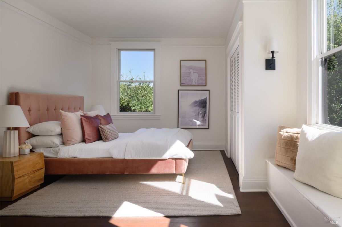 A bedroom with a pink upholstered bed, white walls, and two windows, one with a window seat.