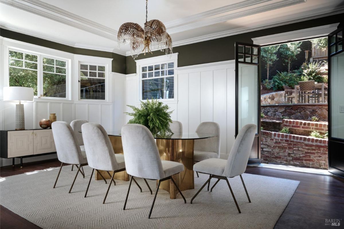 A dining room with dark green walls, white wainscoting, a large window with a view of a patio, and a table with eight chairs.