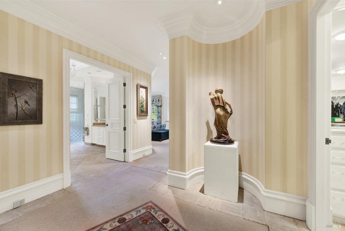 Hallway with striped wallpaper and crown molding features a curved wall showcasing a sculpture and leads to other rooms.