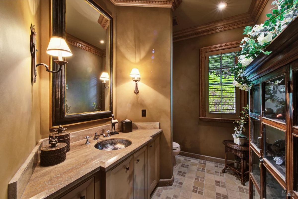 Powder room with a patterned tile floor, a vanity with a marble countertop and a decorative mirror, and a window with plantation shutters.