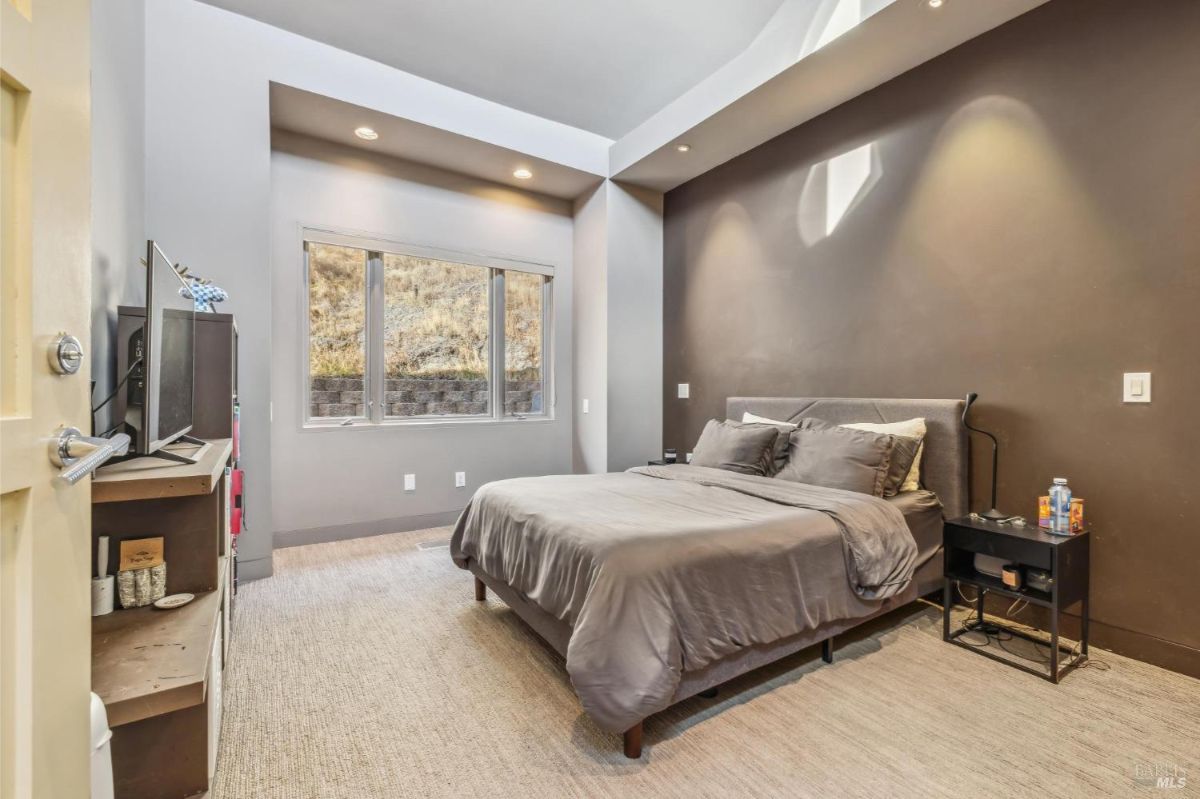 Bedroom with dark accent wall, recessed lighting, and large windows facing a hillside.