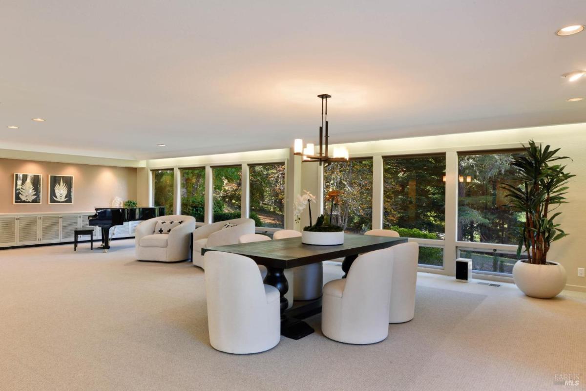 A dining area with white chairs next to large windows overlooking greenery.