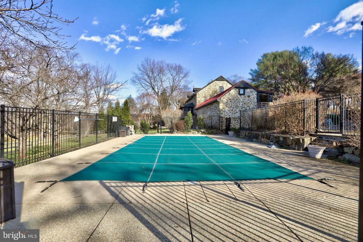A teal pool cover lies over an in-ground rectangular swimming pool, situated behind a black metal fence.