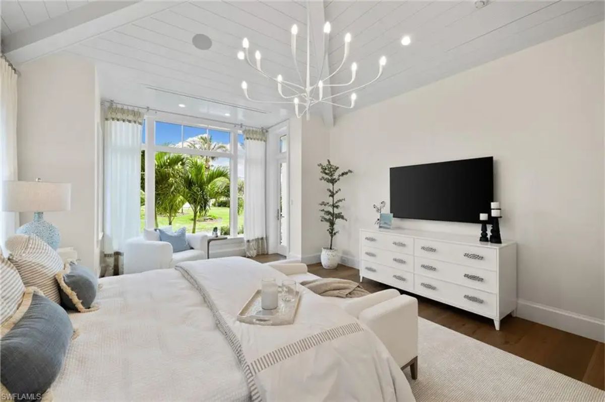 Spacious bedroom includes a white bed, large windows, and a unique chandelier, with views of palm trees and greenery. 