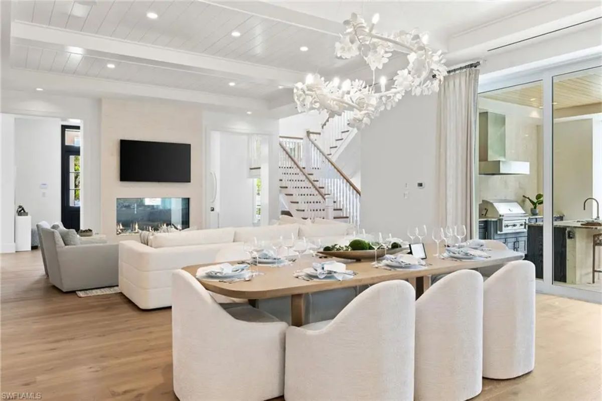 Dining area with a large wooden table, upholstered chairs, and a decorative chandelier near an outdoor kitchen. 