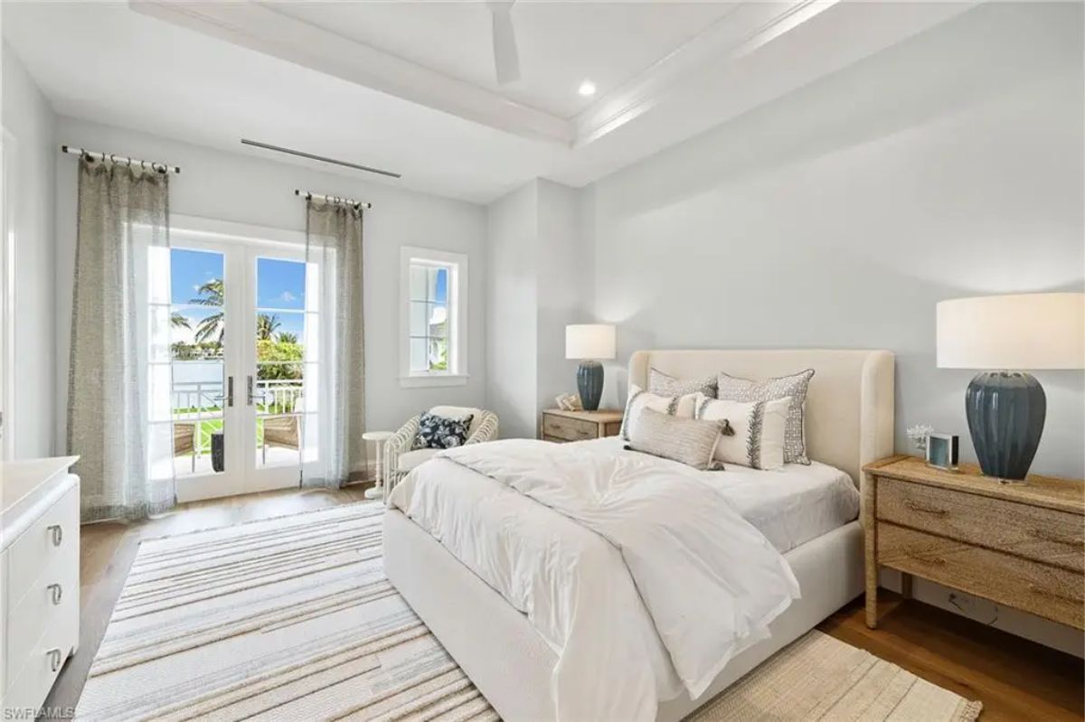 Bright bedroom features a white bed with wooden side tables, large French doors, and striped area rug leading to a balcony. 