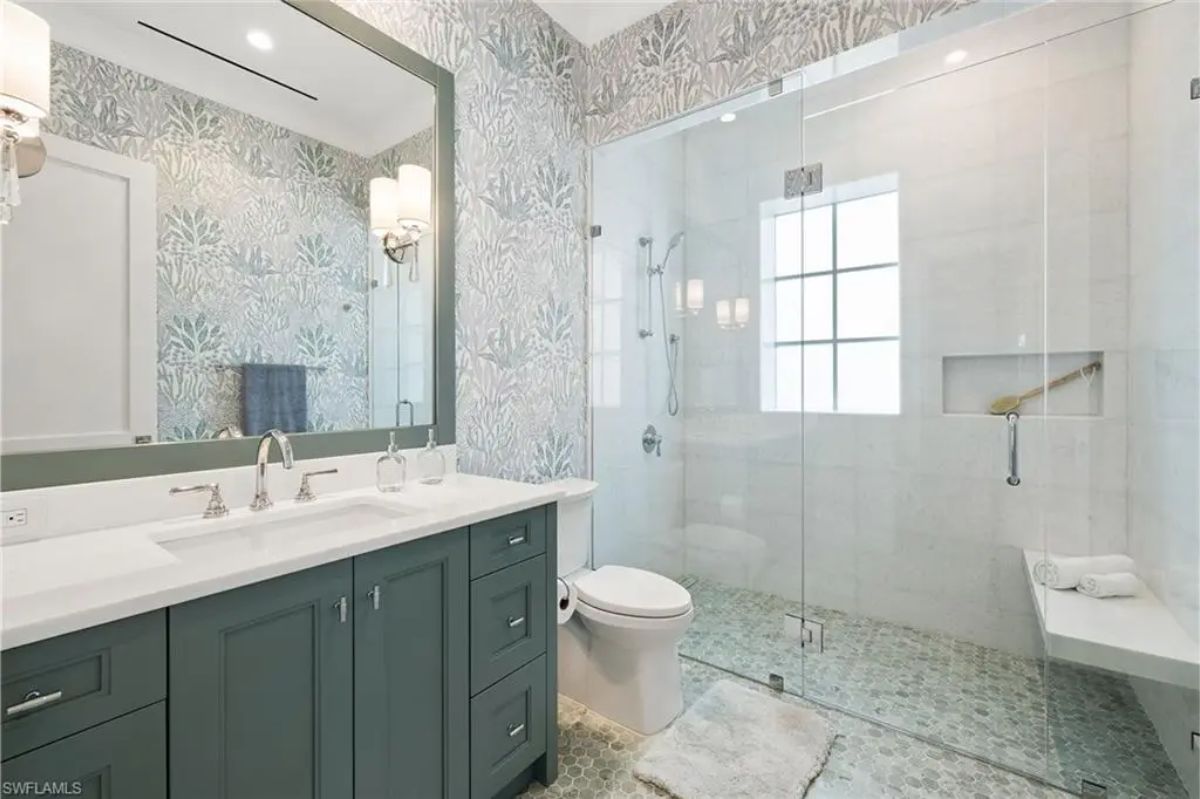 Well-designed bathroom features patterned wallpaper, green cabinetry, and a glass-enclosed walk-in shower. 