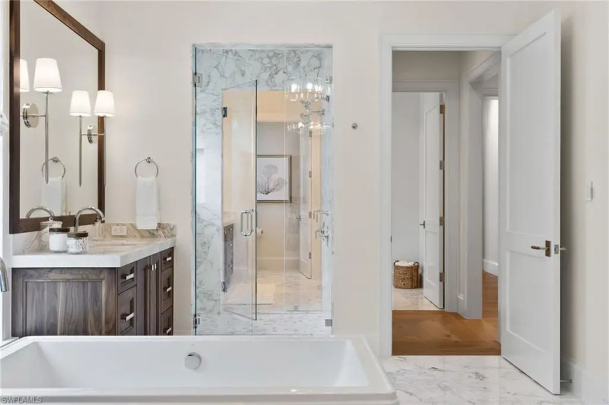 Marble bathroom includes a large soaking tub, glass-enclosed shower, and dual vanities with dark wooden cabinetry. 