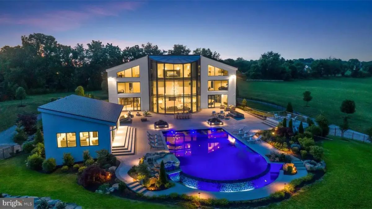 Elevated rear view highlights the illuminated pool area, patio, and expansive lawn at dusk. 