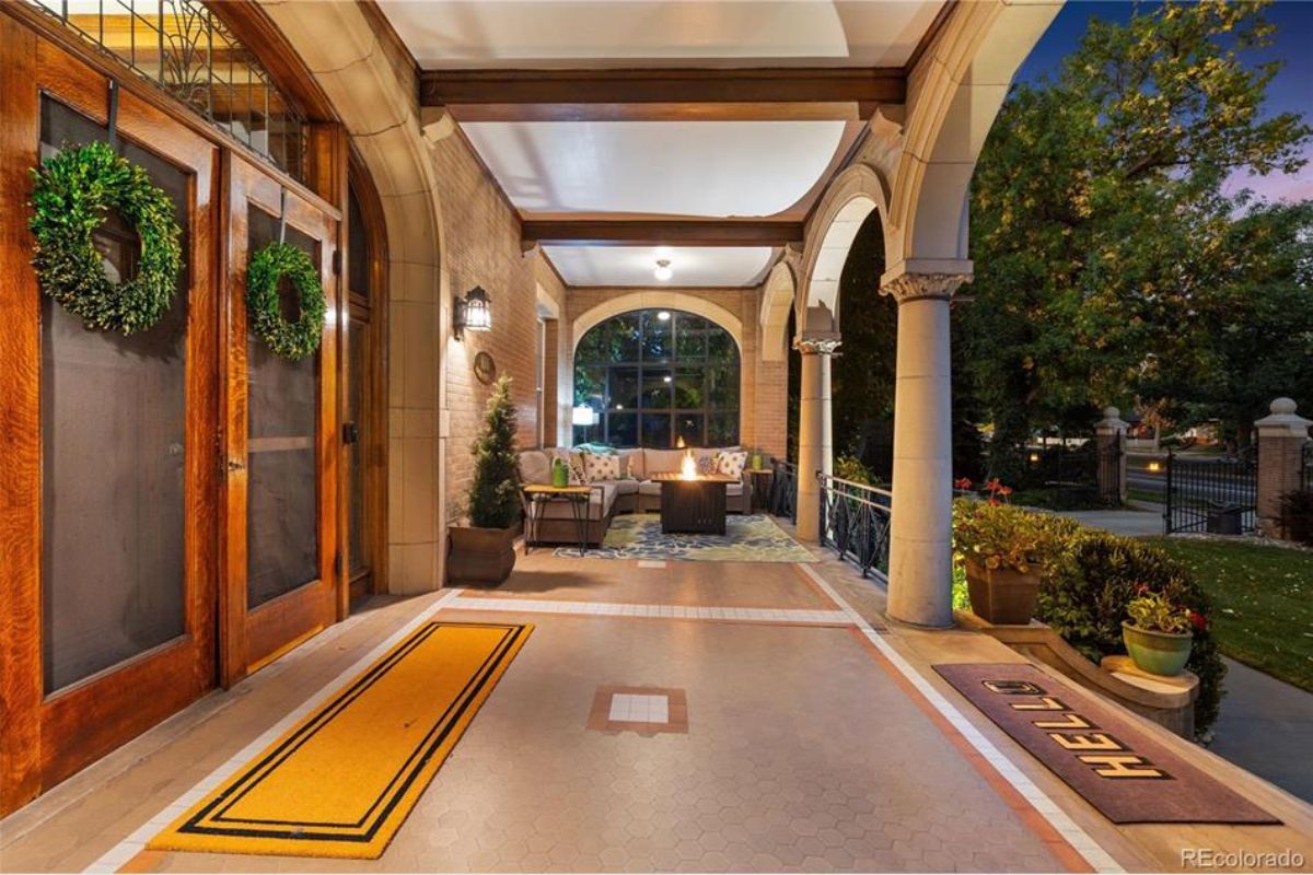 The illuminated porch of the Sharp Mansion showcases its timeless charm with arched columns, intricate tile flooring, and a warm, inviting seating area centered around a fire feature.