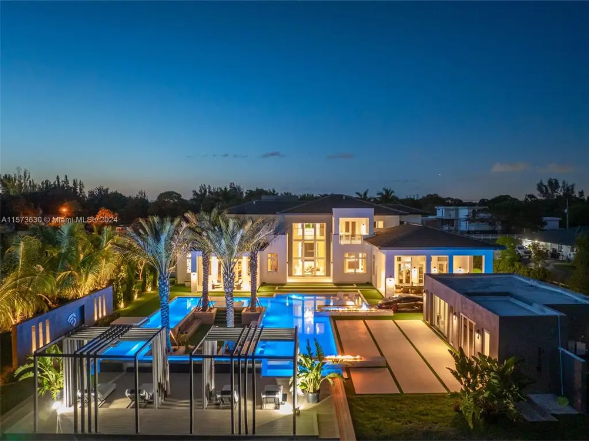 Expansive modern home exterior features a well-lit pool area and palm trees at dusk.