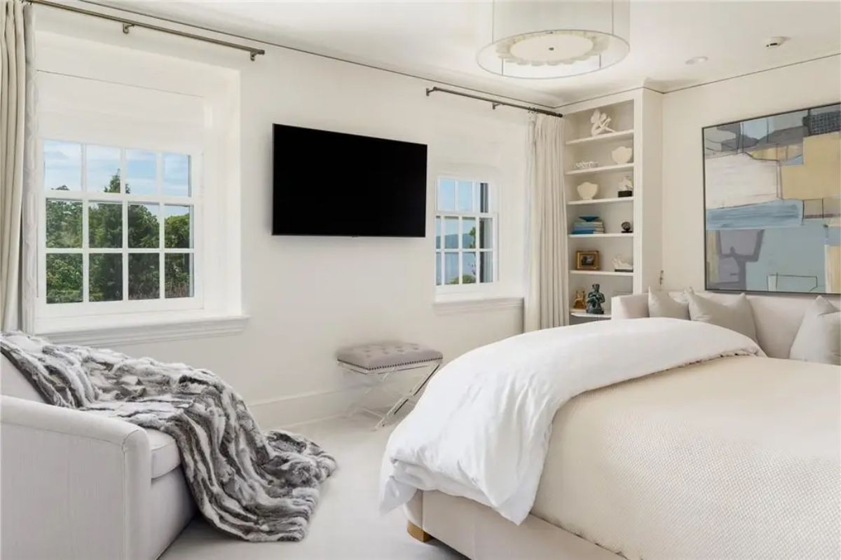 Bedroom with white furnishings, built-in shelving, and large windows providing natural light. 