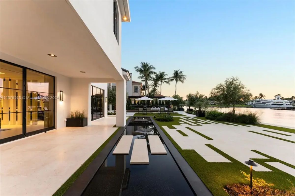 Outdoor patio area showcases a modern landscape design with geometric grass patterns, a reflection pond with floating steps, and a view of the waterfront. 
