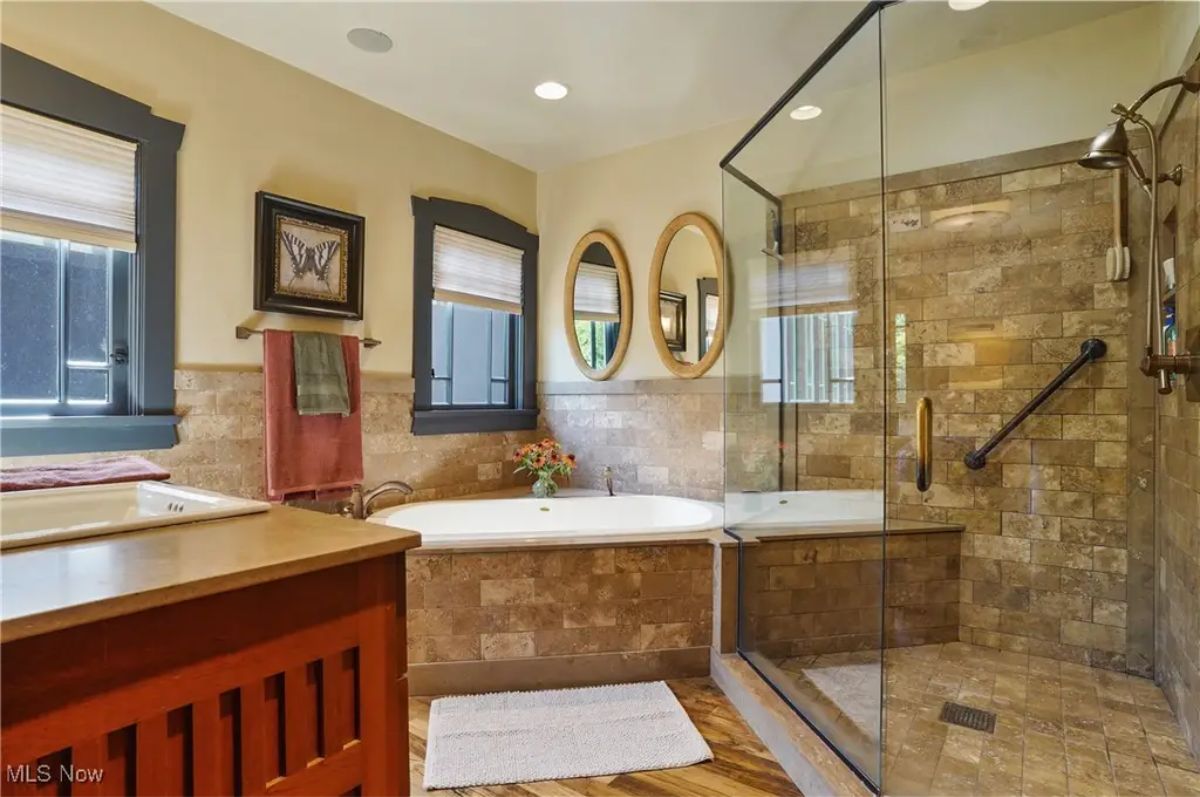Soaking tub framed by neutral stone tile and complemented by a large glass-enclosed shower.