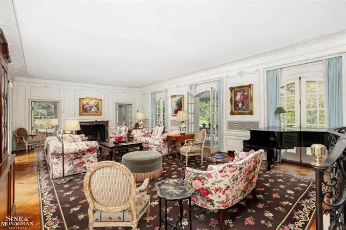 Spacious living room with white-paneled walls and detailed moldings. Large windows and French doors allow natural light to illuminate the space.
