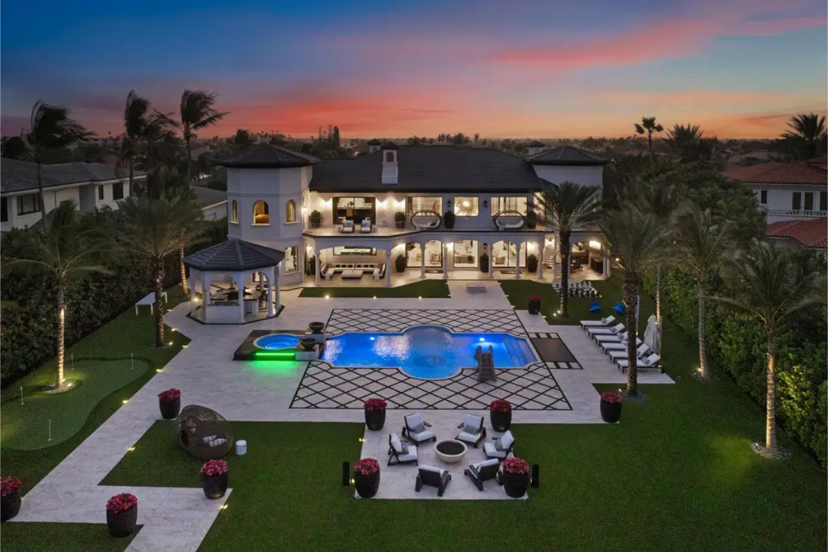 Grand estate at dusk with an illuminated pool, gazebo, and a landscaped backyard featuring lounge areas and a fire pit. 