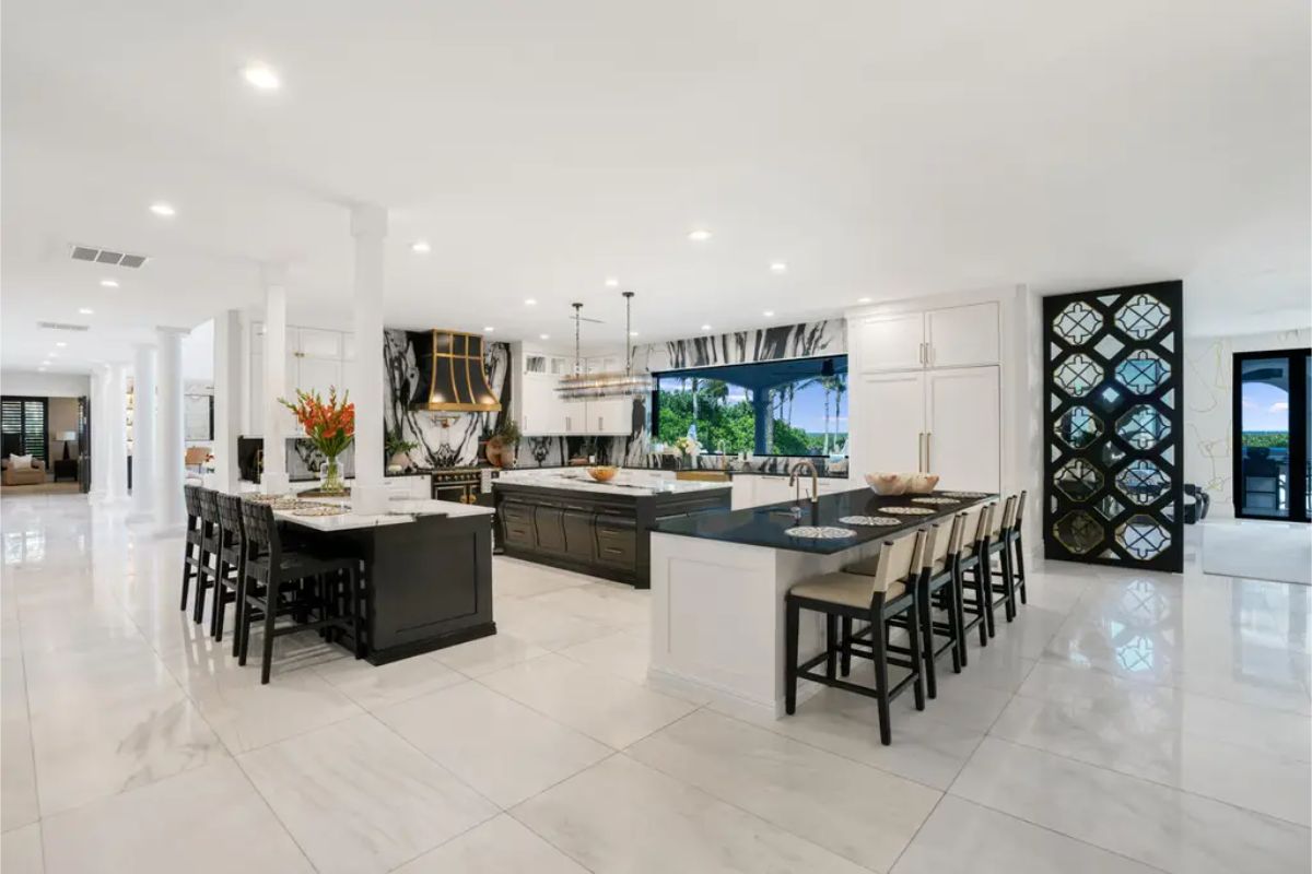Kitchen with white marble floors, two kitchen islands with seating, white cabinets, and large windows overlooking a green outdoor area. 