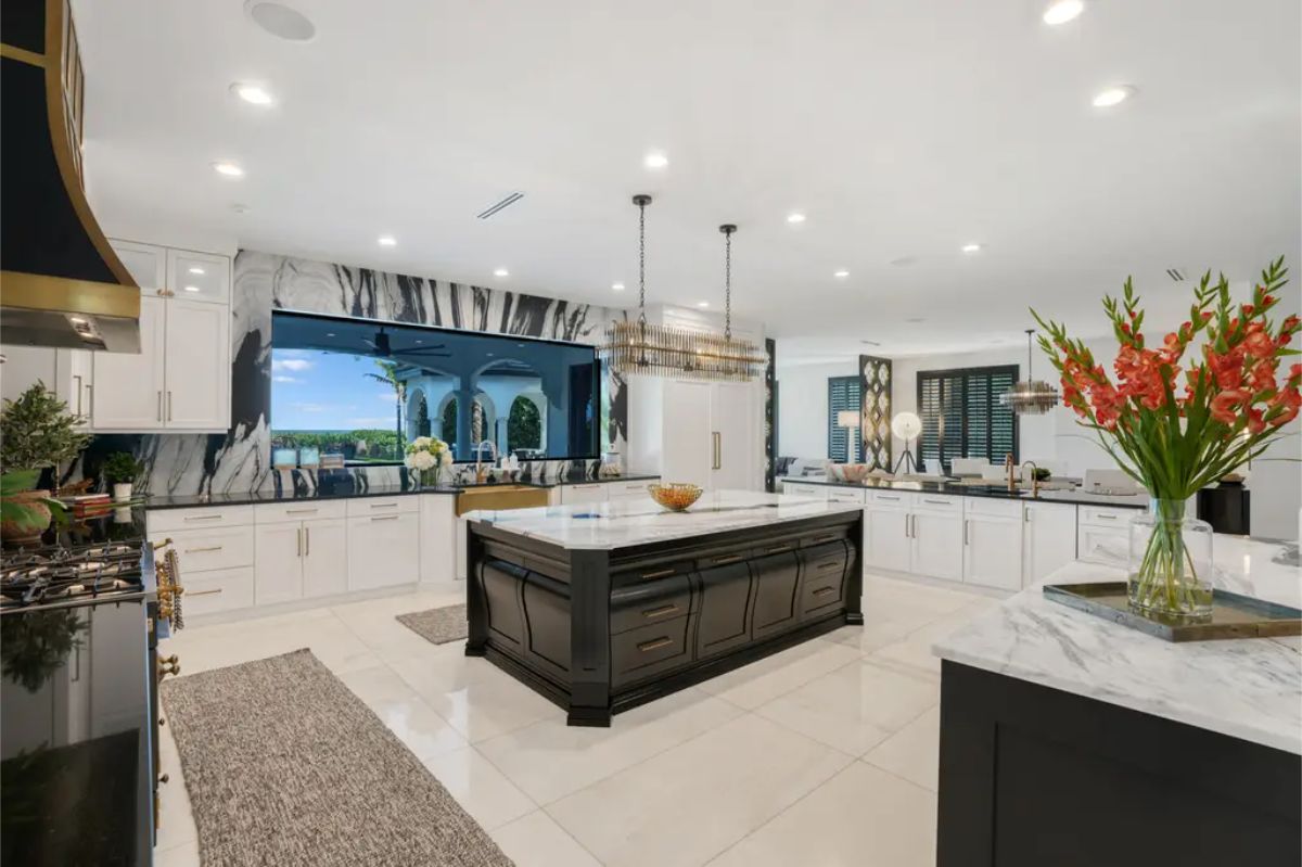 Kitchen with white cabinets, a large black kitchen island with a white countertop, a marble backsplash and range hood, and a large window overlooking an outdoor scene.
