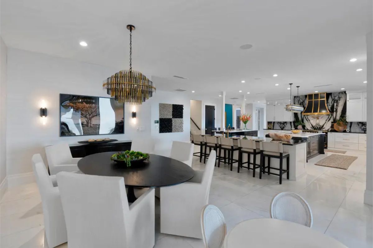 A modern dining area with a round black table and white upholstered chairs sits adjacent to a contemporary kitchen featuring a large island with seating, white cabinetry, and a decorative range hood.