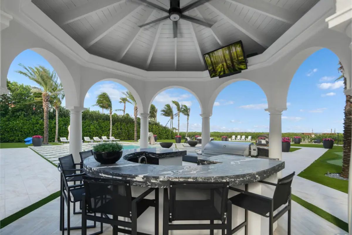 Outdoor bar area under a gazebo with marble countertops, black chairs, and a built-in grill, overlooking the lush backyard. 