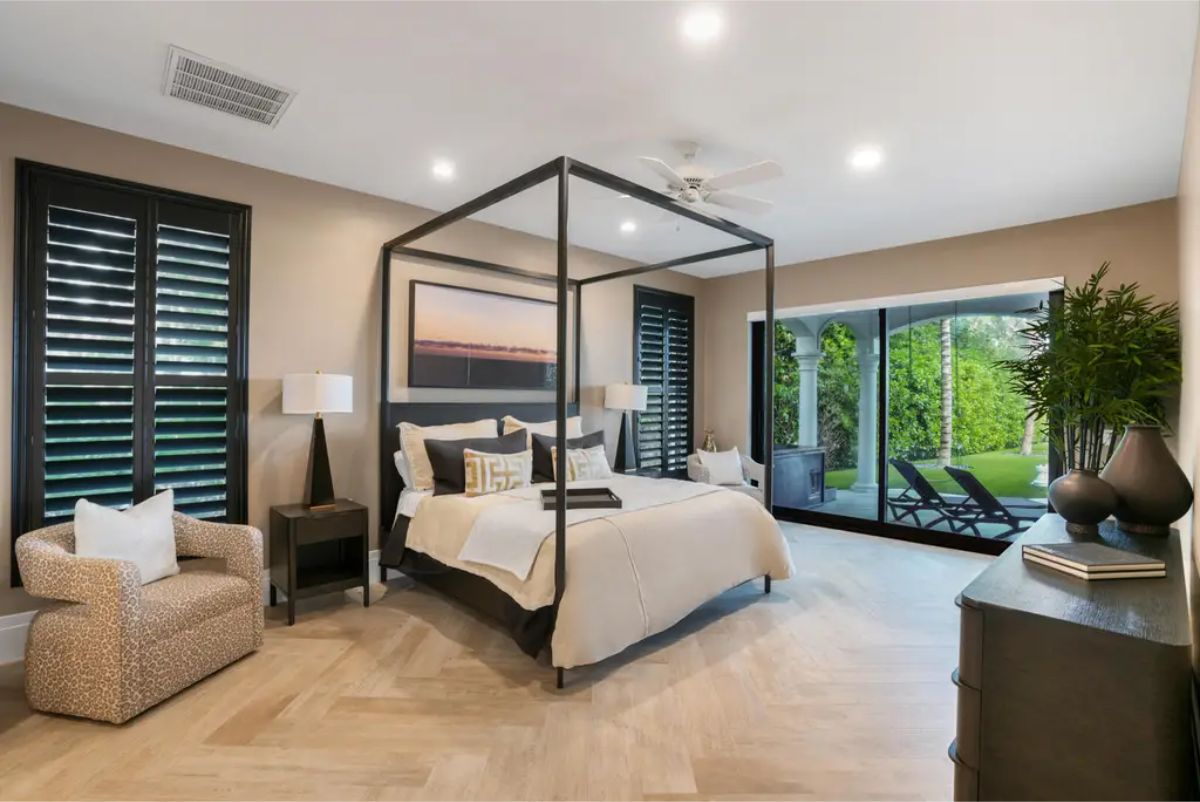 Bedroom boasts a four-poster bed with a dark frame, complemented by neutral tones in the bedding and walls, and features large windows overlooking a lush outdoor patio.