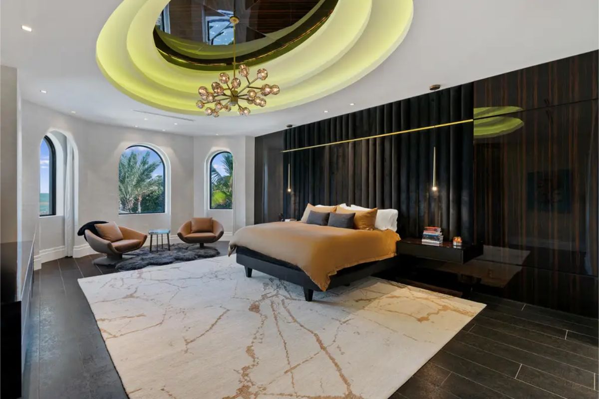 Unique, layered ceiling design with a central light fixture, a dark accent wall with built-in lighting, and arched windows overlooking a lush landscape. 