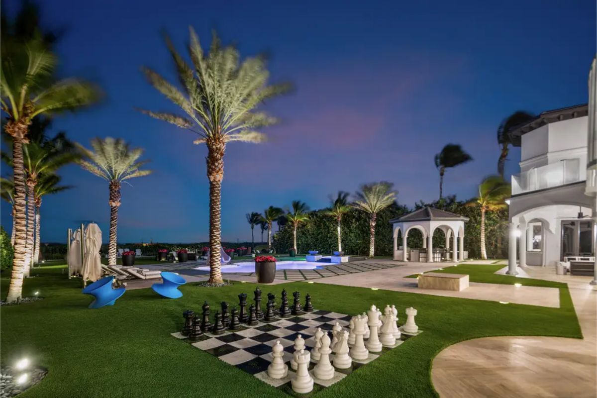 Nighttime backyard scene with illuminated palm trees, a giant outdoor chess set, and a modern pool area. 