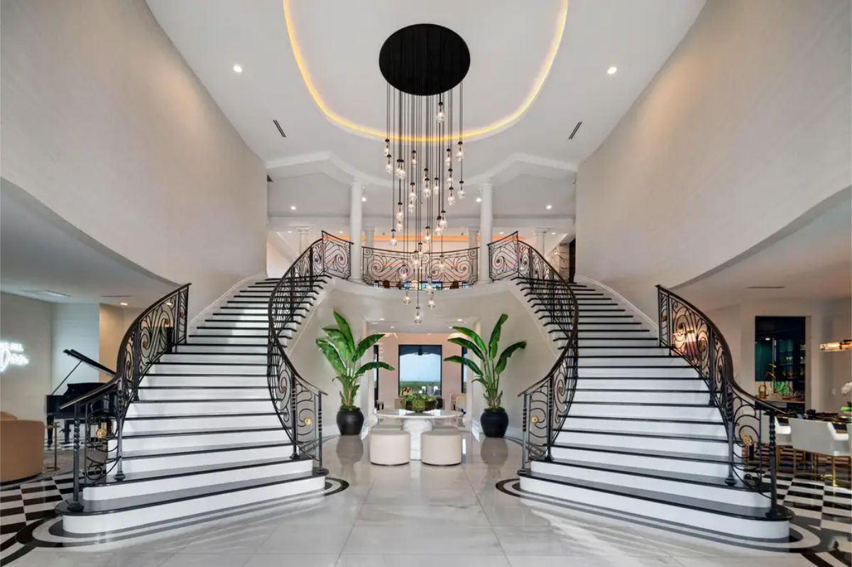 Grand foyer featuring a double staircase with intricate black railings, white marble floors, and a modern chandelier. 