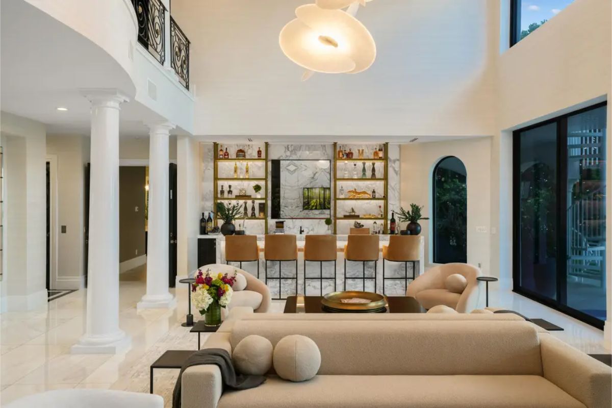 Living room with tall ceilings, white walls, a prominent central light fixture, a bar area with stools, and large windows.