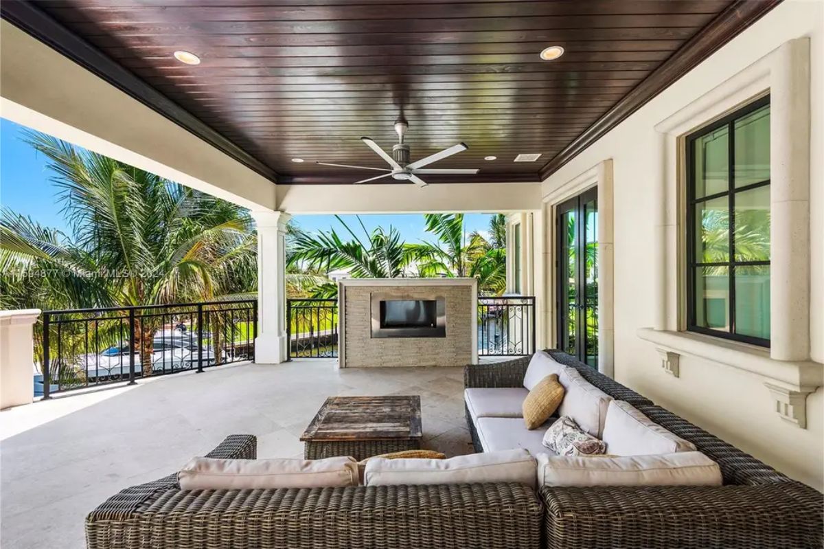 Covered outdoor seating area with a ceiling fan and built-in fireplace overlooking lush palm trees. 